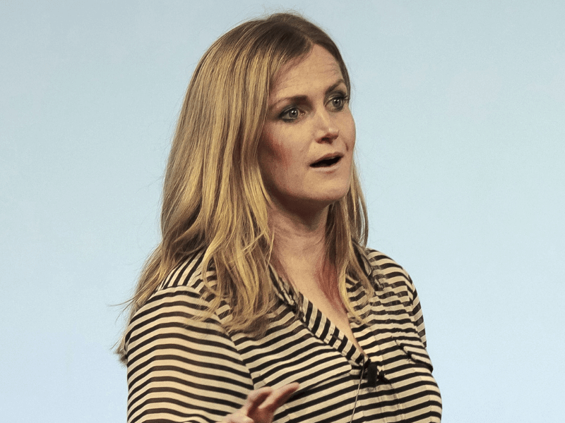 Katherine Trebeck speaking confidently on stage, wearing a striped shirt and gesturing to emphasize her point